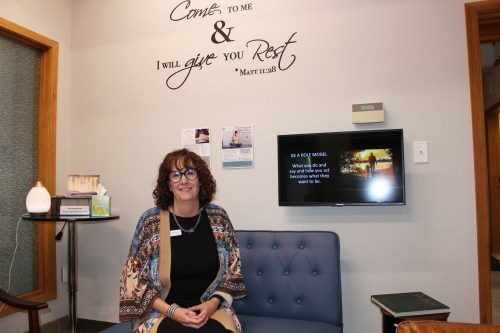 A woman sits in a chair at a crisis pregnant center. The wall behind her has Biblical scripture written on it: "Come to me & I will give you rest." A TV monitor next to her shows a slide that reads, "Be a role model. What you do and say and how you act becomes what they want to be."