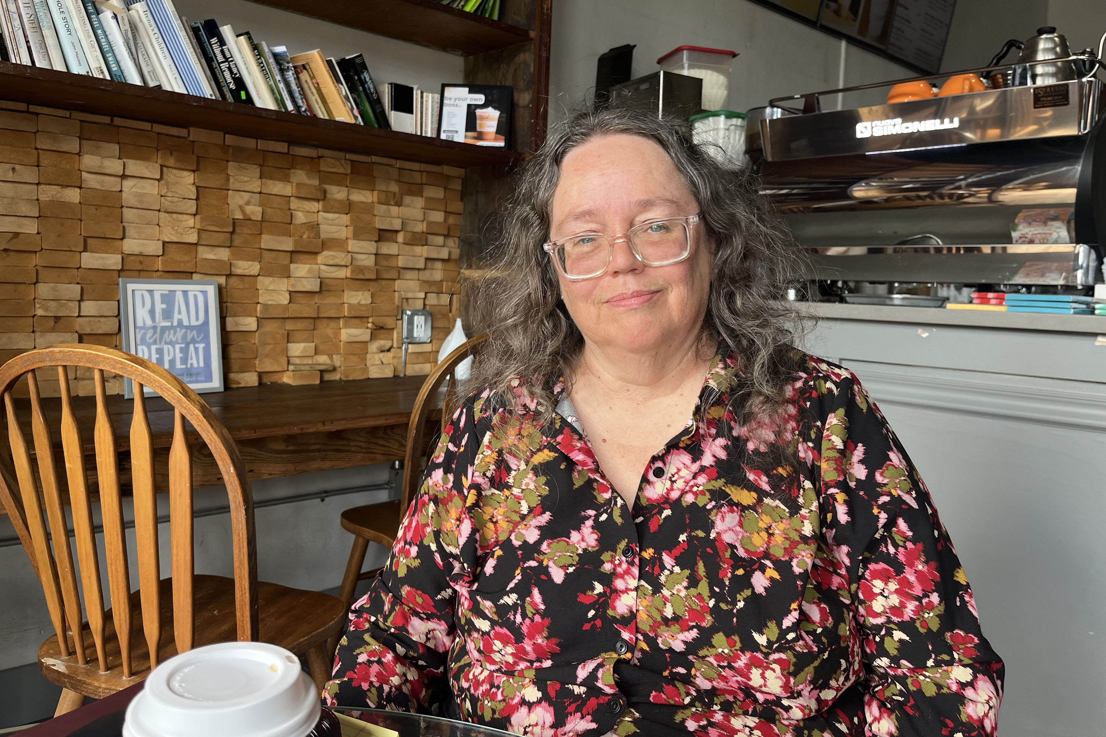 A woman with long, wavy gray hair, wearing a floral blouse and glasses, sits at a table in a coffee shop. She looks in the direction of the camera with a calm expression.
