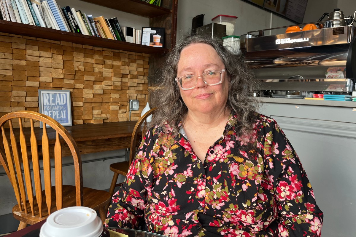 A woman with long, wavy gray hair, wearing a floral blouse and glasses, sits at a table in a coffee shop. She looks in the direction of the camera with a calm expression.