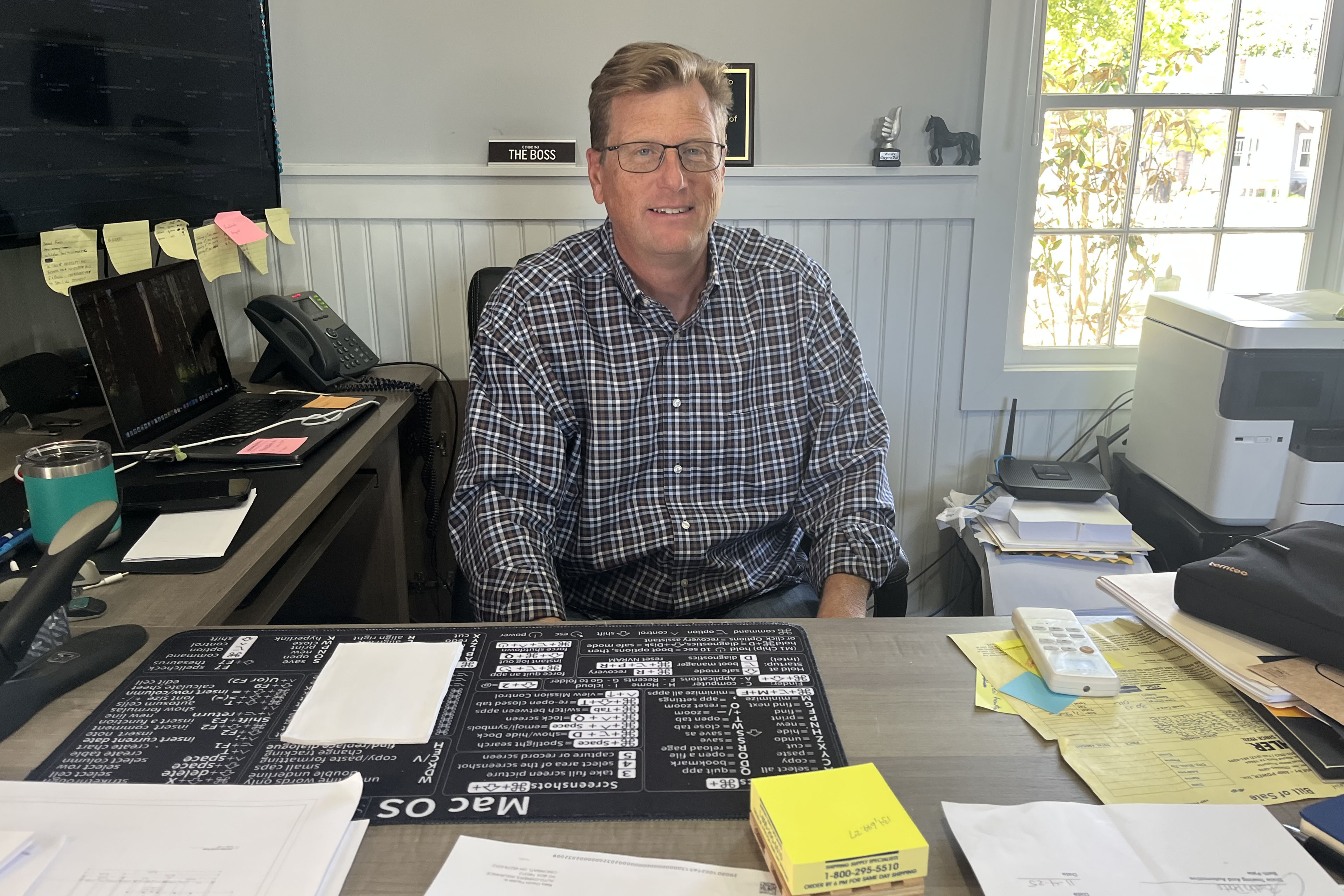 A man with short brown hair wearing a button down shirt and glasses sits at an office desk. He is surrounded by two computers, papers and post-it notes, and a printer.