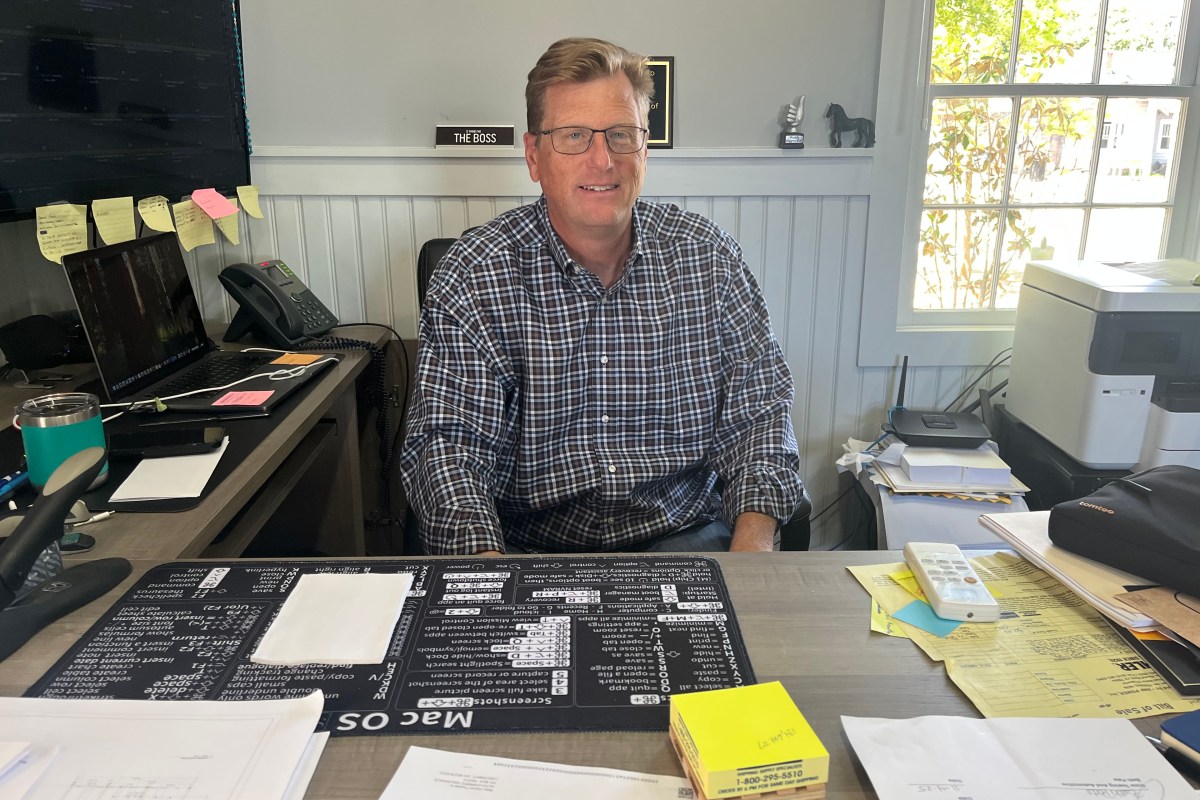 A man with short brown hair wearing a button down shirt and glasses sits at an office desk. He is surrounded by two computers, papers and post-it notes, and a printer.