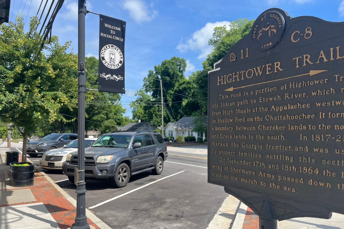 A photo shows an outdoor parking area of a small town. A sign on a lamp post reads, "welcome to Social Circle." A historic sign in the foreground tells the history of the Hightower Trail.