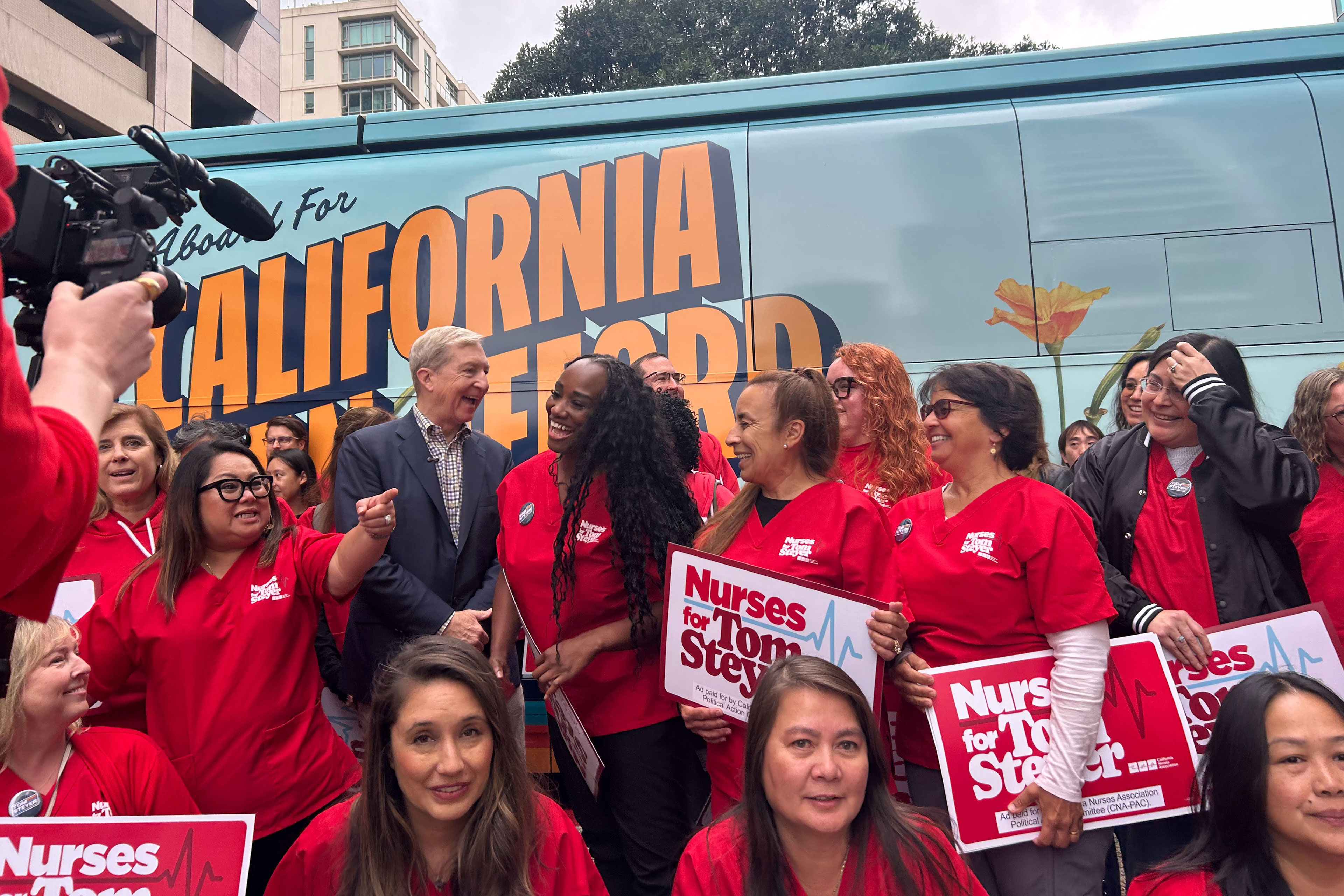 A group of people — most wearing red shirts — mingle in front of a bus with the words "All Aboard For A California You Can Afford" on its side.