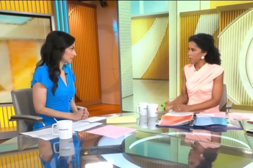 Two women behind a desk on a TV news set speak to each other. Papers and mugs are on the desk.