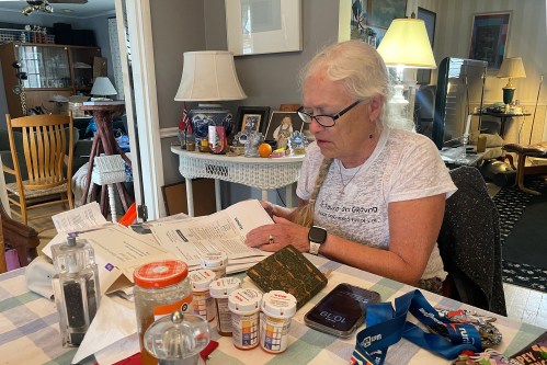 A woman with white hair in a braid sits at a dining room table and looks through paperwork. A notebook, pill bottles, a phone, and a medal on a ribbon are on the table in front of her.