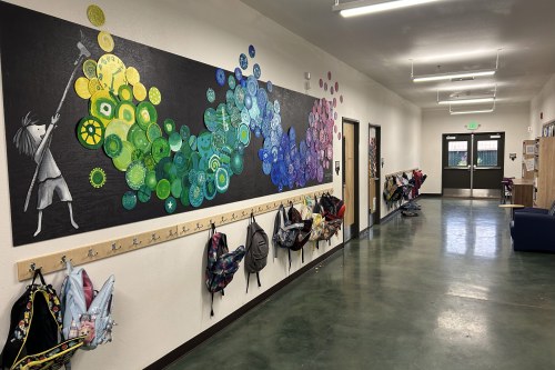 A photo of a hallway in a school. A black panel lines the wall with an illustration of a child painting a cascading gradient from yellow, blue to purple. Backpacks hang from hooks underneath it.