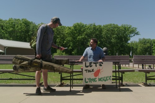 A reporter seated at a makeshift table outside. A man on the left approaches to speak with him. A sign in front of the table reads, "Let's talk Lyme disease."