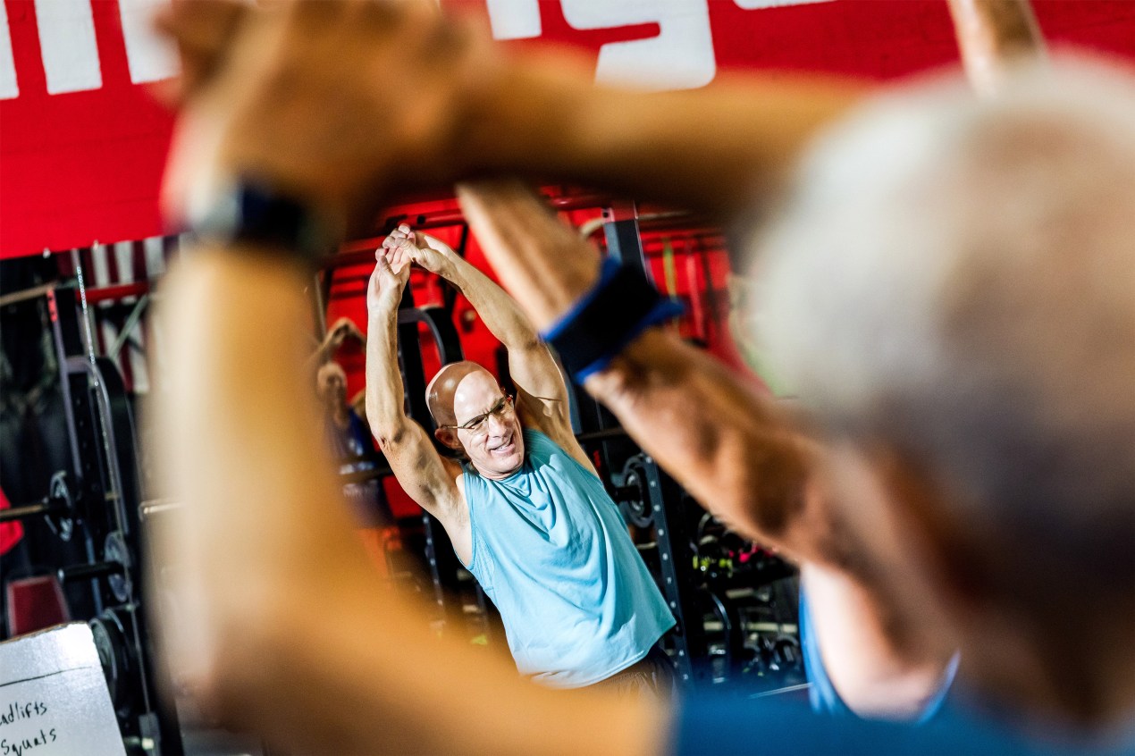 Larry Gruber stretches both arms upward, hands clasped together. A blurred figure in the foreground does the same stretch, framing his face.
