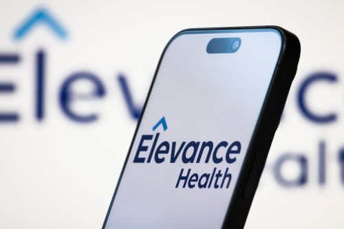 A photo illustration shows Elevance Health's logo on a phone screen and on a monitor behind the phone.