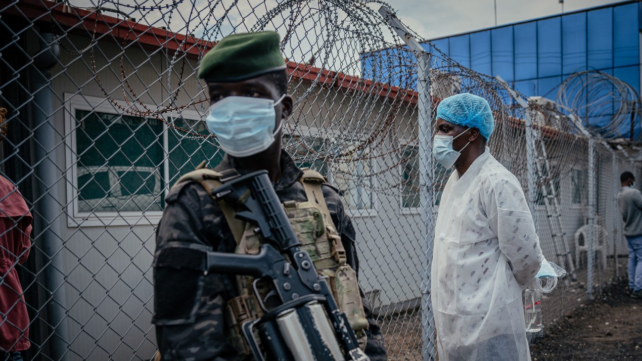 An armed guard stands in front of a building behind a barbed wire fence. A medical worker wearing a gauze cap, mask, and gown, looks at the building.