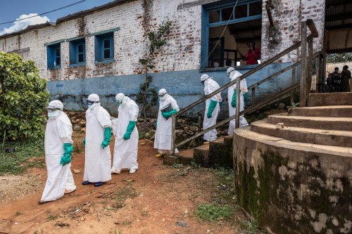 A line of medical workers in Congo walk out of a building in protective gear: caps, goggles, face masks, long white body coverings, and teal gloves.