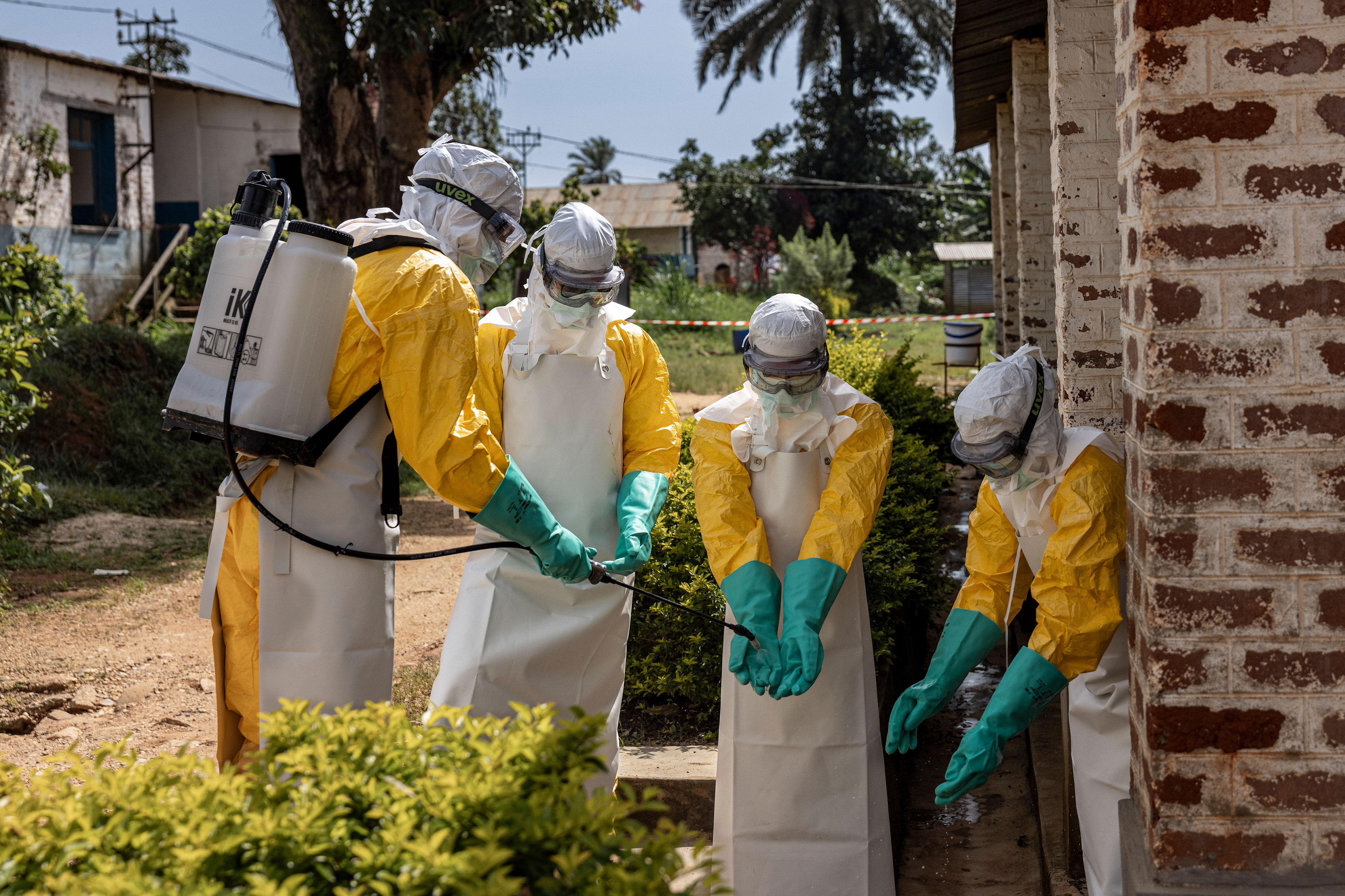 A person wearing tank on their back holds out a hose to dispense liquid from it onto the gloved hands of three other people. All four people wear personal protective equipment from head to toe, including goggles and head coverings.