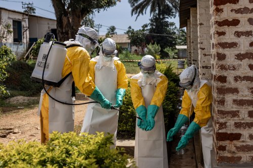 A person wearing tank on their back holds out a hose to dispense liquid from it onto the gloved hands of three other people. All four people wear personal protective equipment from head to toe, including goggles and head coverings.