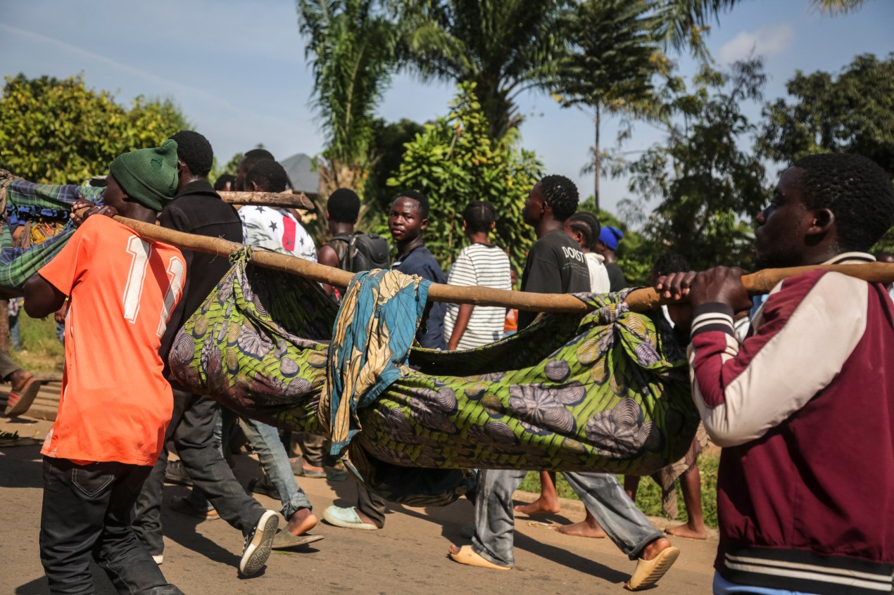 Two people transport a body cradled in a cloth alongside a group of people walking.