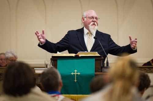A reverend speaks at a lectern. People are seated in front of him, blurred in the foreground.