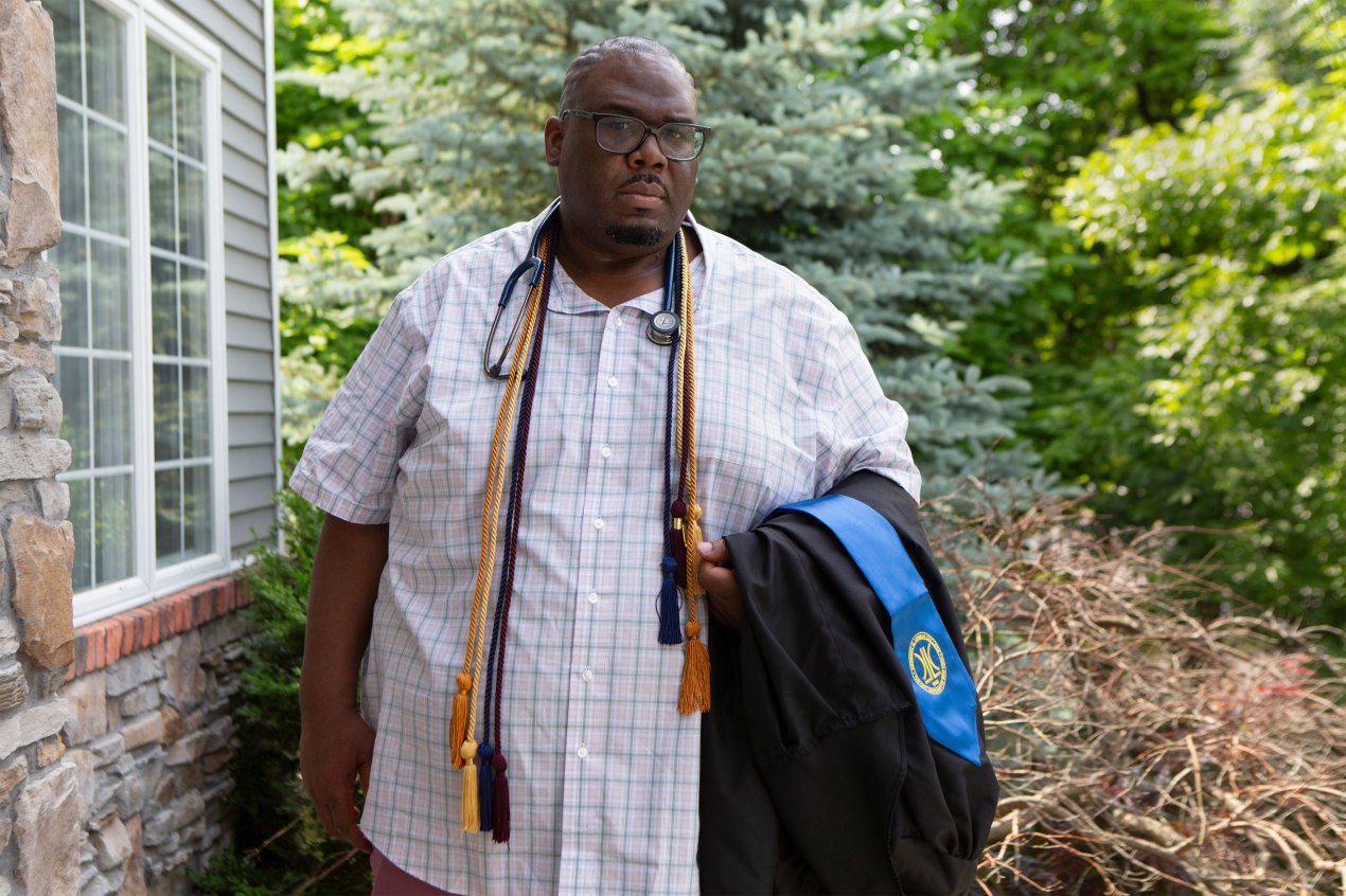 Benjamin Pinckney stands outside. He is holding his graduation gown and has his graduation cords draped over his neck.
