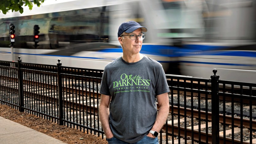 A man wearing a navy baseball cap and a gray shirt that reads "Out of the Darkness community walks" stands along light rail tracks with a train passing behind him