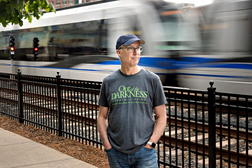 A man wearing a navy baseball cap and a gray shirt that reads "Out of the Darkness community walks" stands along light rail tracks with a train passing behind him