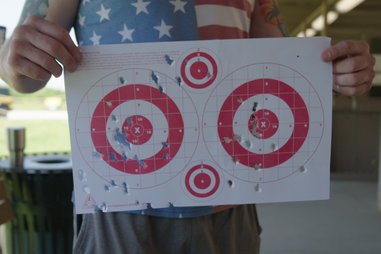 A pair of hands holds up a piece of paper with two large bull's eye targets and two small ones. Bullet holes are scattered across the paper.
