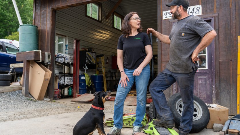 A woman and man stand by an auto shop building. A dog sits in front of them.