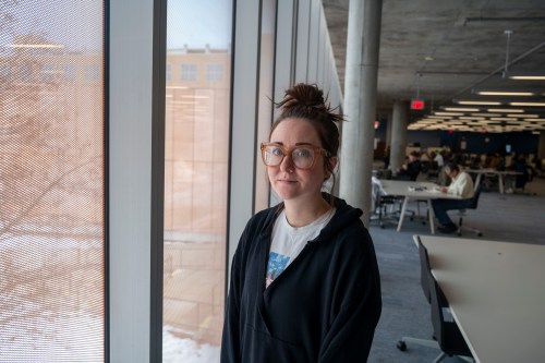 A woman with brown hair tied up in a bun stands inside a building near some windows. Snow is on the ground outside and people sit a large tables behind her.