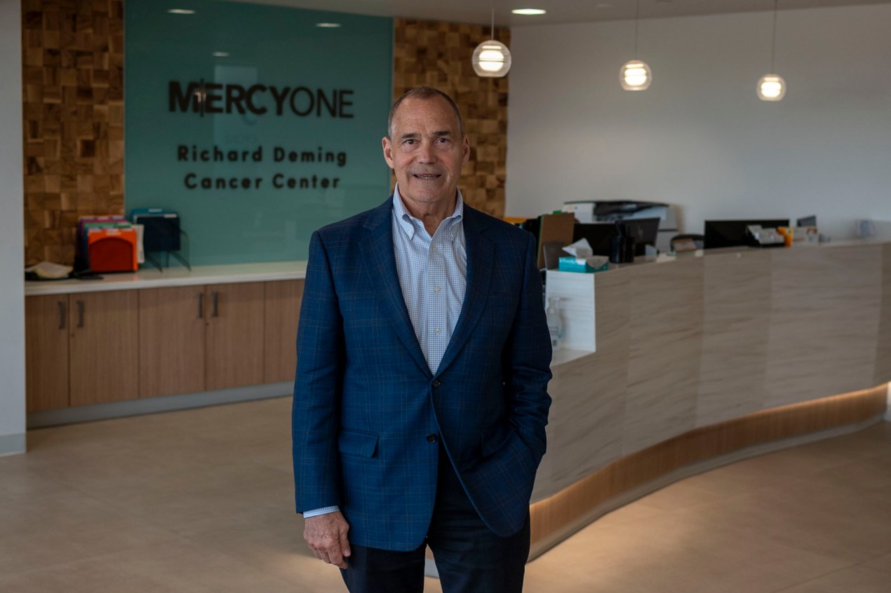 A man in a dark blue suit jacket stands in front of a reception desk. A sign reading "MercyOne Richard Deming Cancer Center" is on the wall behind the desk.