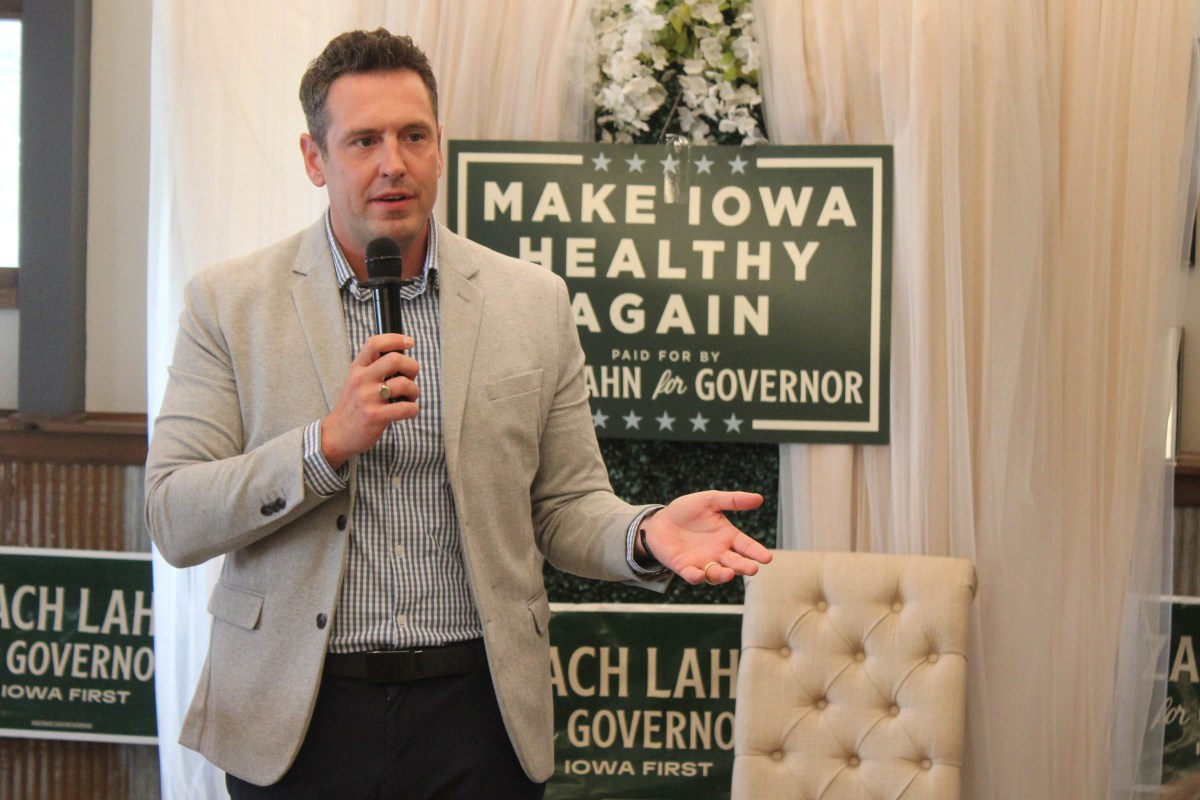 A man in light colored blazer and button down shirt holds a microphone. Behind him, campaign signs that say "MAKE IOWA HEALTHY AGAIN" are visible.