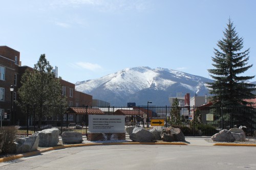 The metal gate with a sign that reads "Rocky Mountain Laboratories" framed by pine trees and snow capped mountain in the background