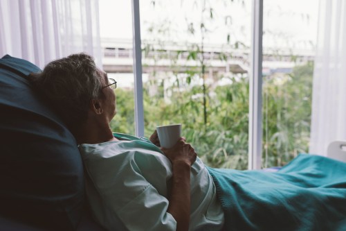 A senior man is lying in a hospital bed while holding a mug and looking out a large window.