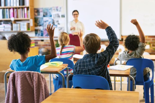 A photo of elementary school students seated at desks, raising their hands to answer a teacher's question. The female teacher is blurred in the background, standing by a whiteboard, calling on a student.