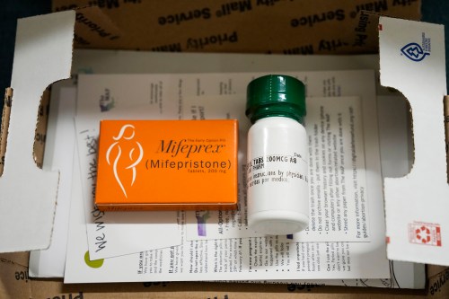 An orange box of mifepristone is beside a white, sealed pill bottle. There are papers underneath, contained in a small cardboard box.