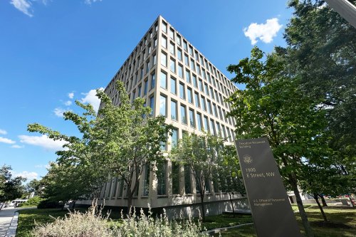 A picture of a building from a low angle. There are trees and a sign that reads "Theodore Roosevelt Building, 1900 E Street, NW, U.S. Office of Personnel Management."