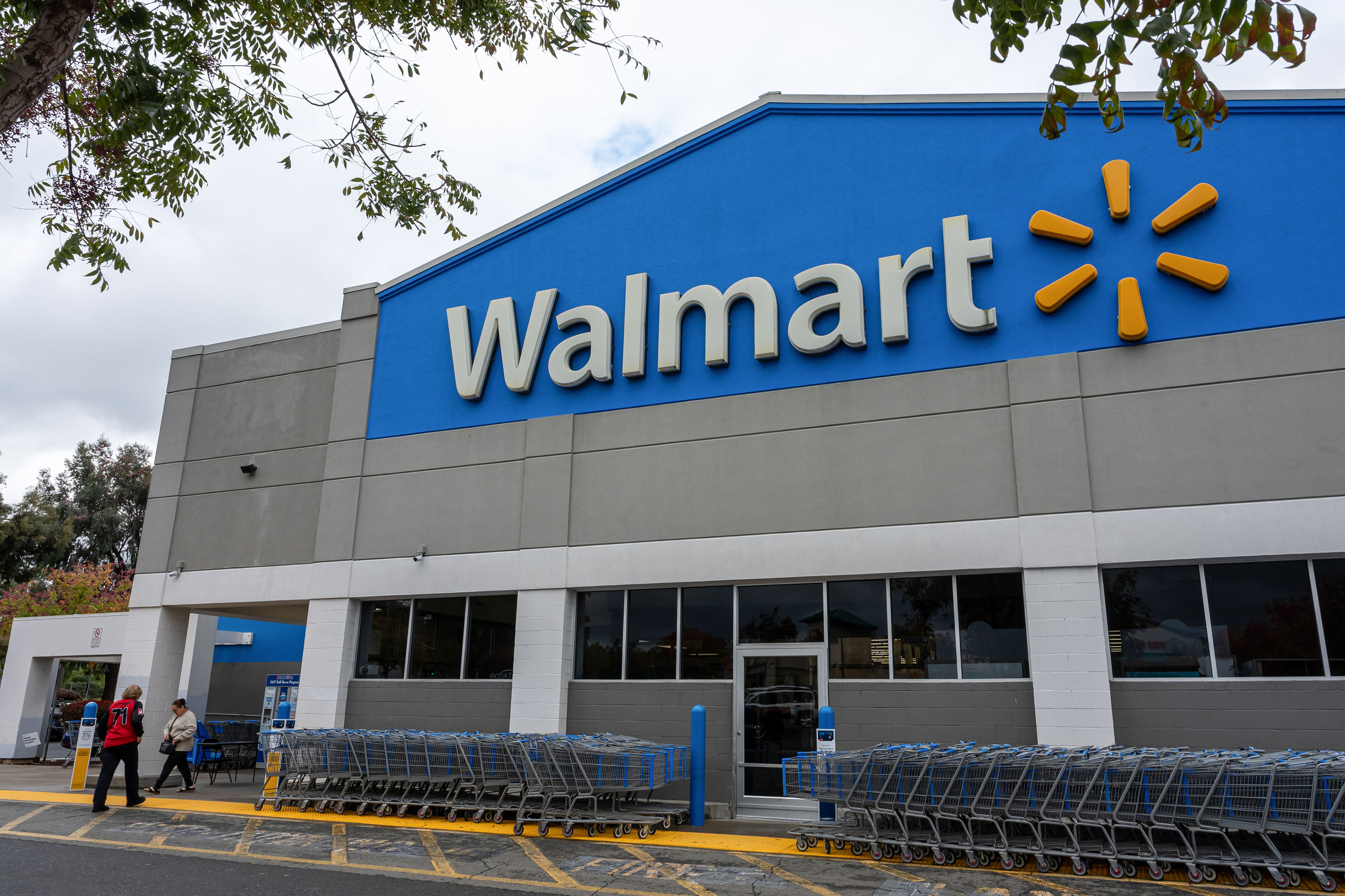 An exterior of a Walmart store. It is a large gray building with Walmart's blue logo covering the top half. Rows of shopping carts line the entryway.