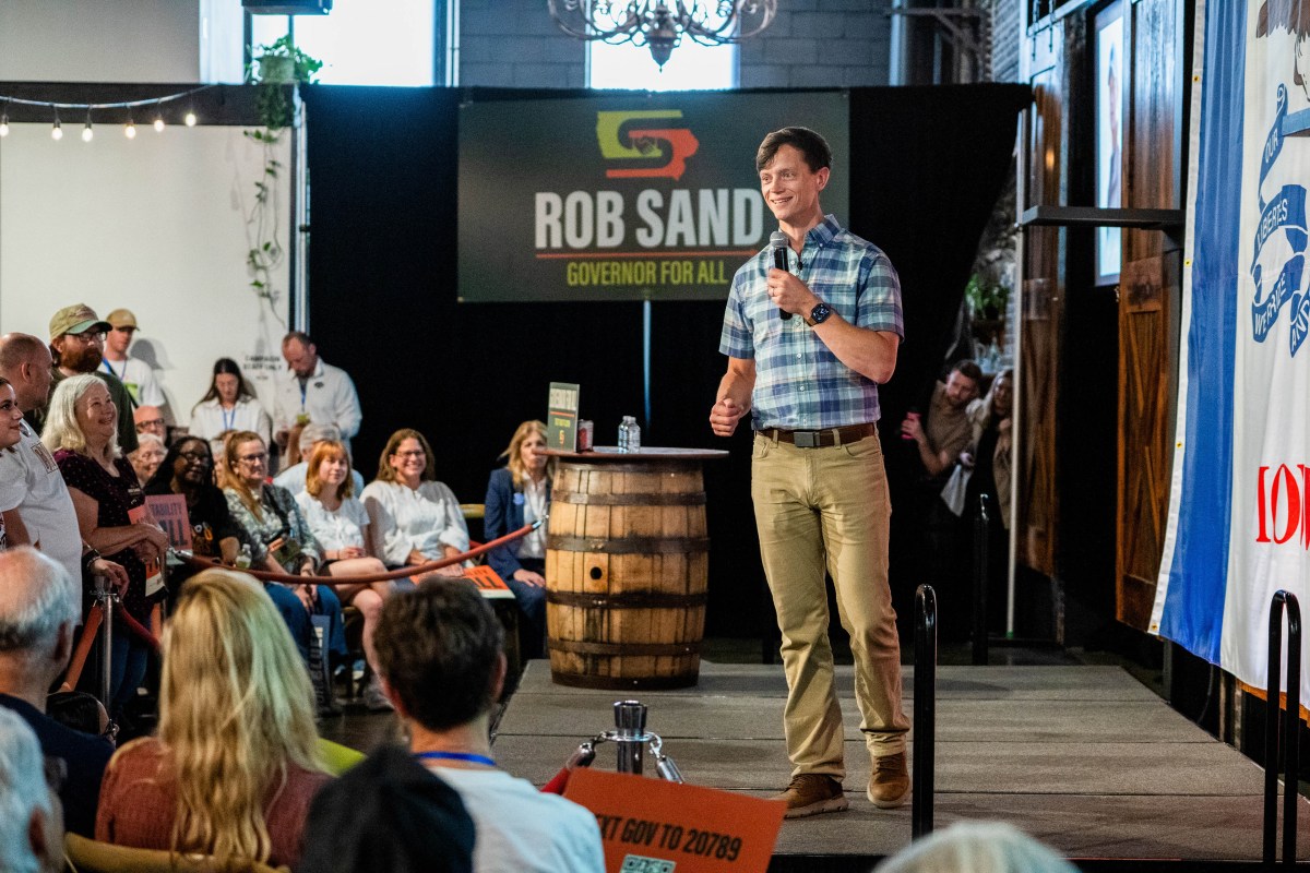 A man wearing a button down shirt and beige pants holds a microphone as he speaks to a small crowd of people.