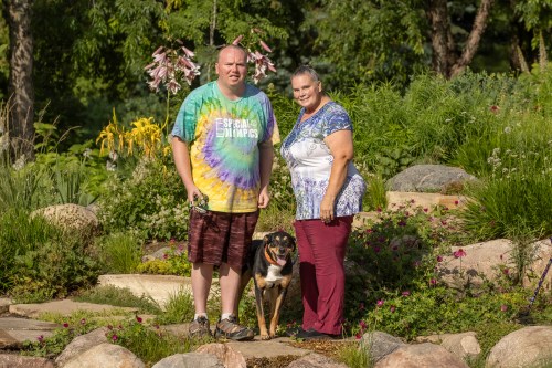 A man and woman stand outdoors with a service dog between them.