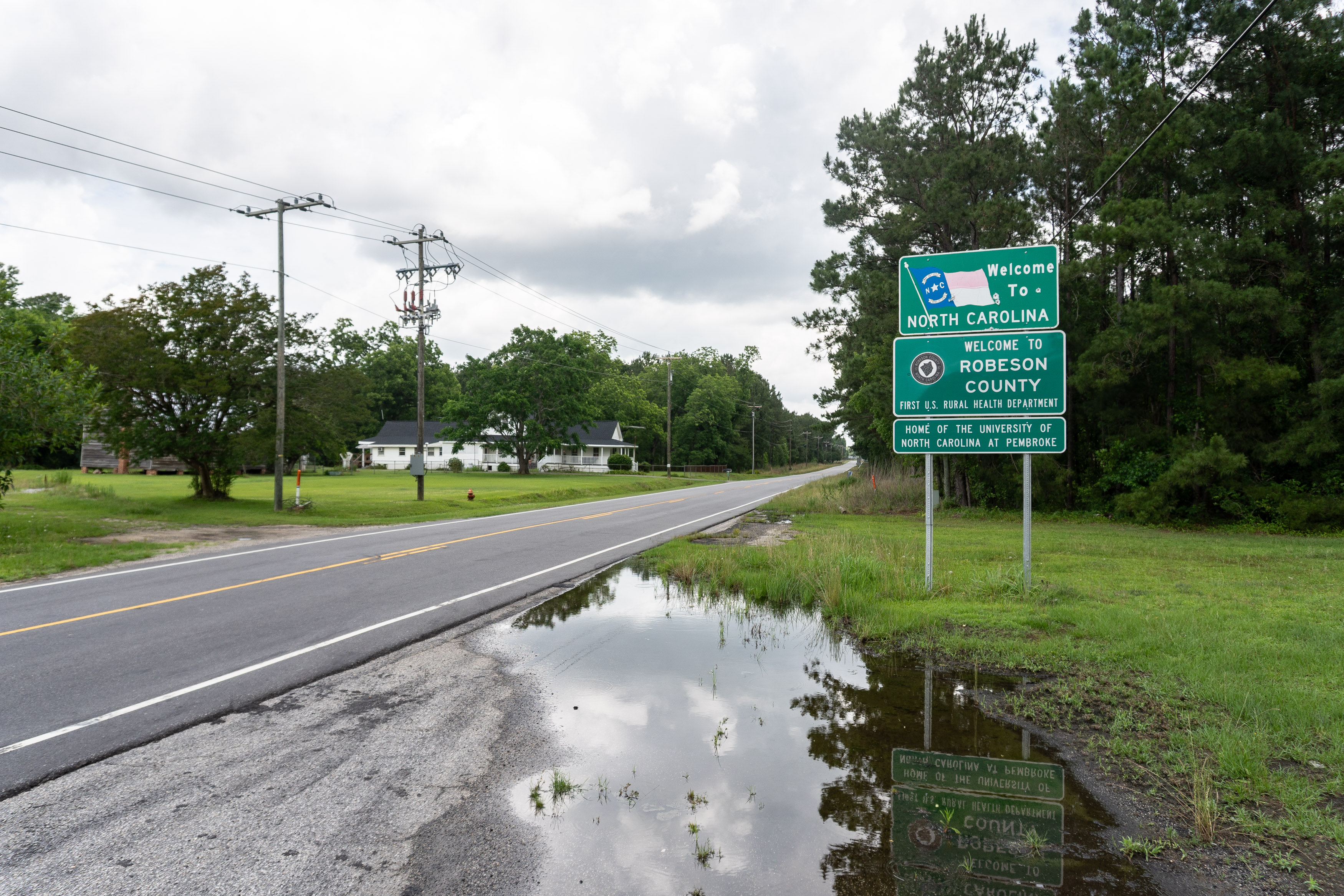 A photo taken from the side of the road shows three signs. They read: "Welcome to North Carolina." "Welcome to Robeson County; first U.S. rural health department." "Home of the University of North Carolina at Pembroke."