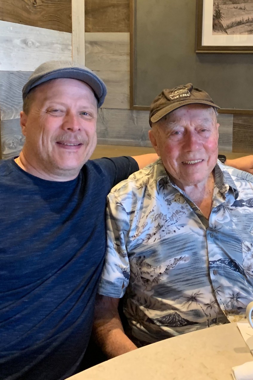 Two men, sitting at a table and wearing hats, smile as their picture is taken