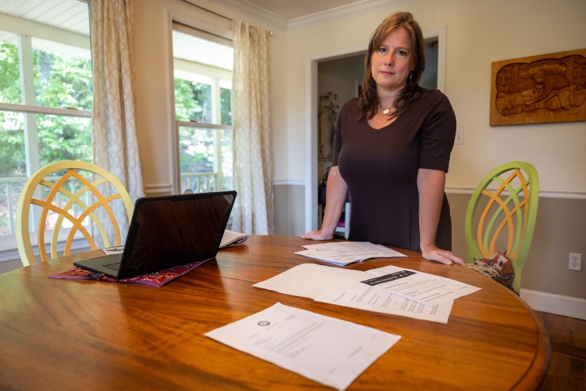 A woman in a brown dress leans on a table with paperwork and a laptop computer in front of her