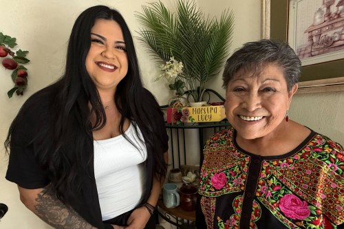 A woman with long brown hair stands beside a senior woman with short gray hair. Both are smiling happily towards the camera.