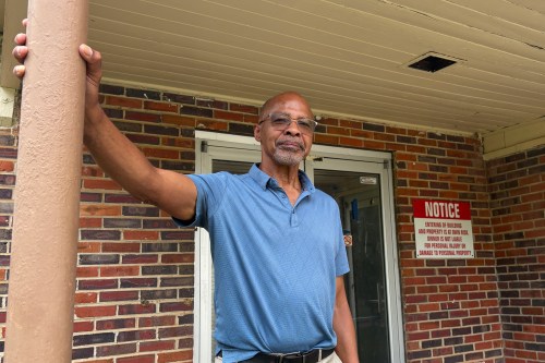 A Black man stands on a porch of a brick building.