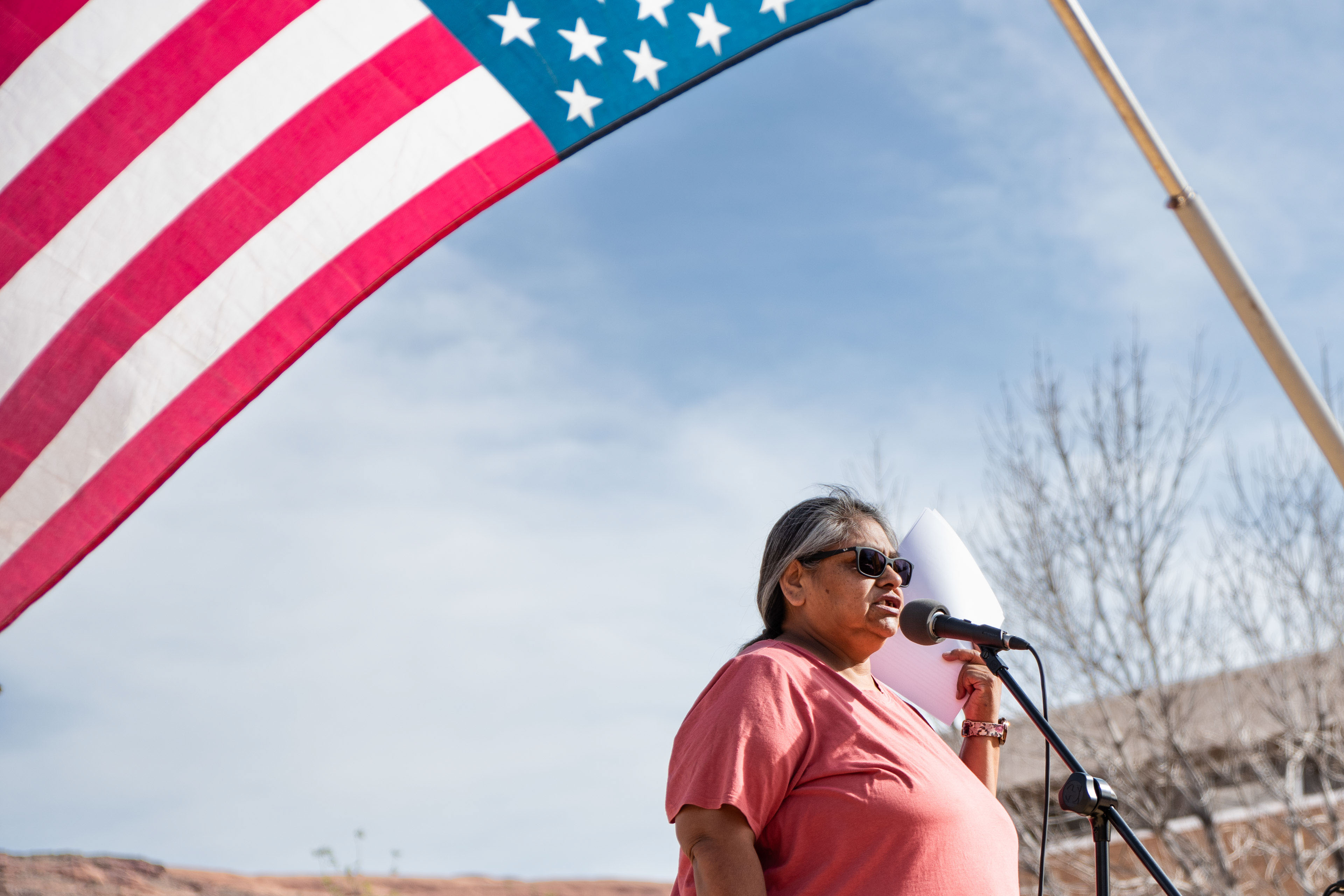 A Native American woman speaks at a microphone. An American flag is seen flying above her.