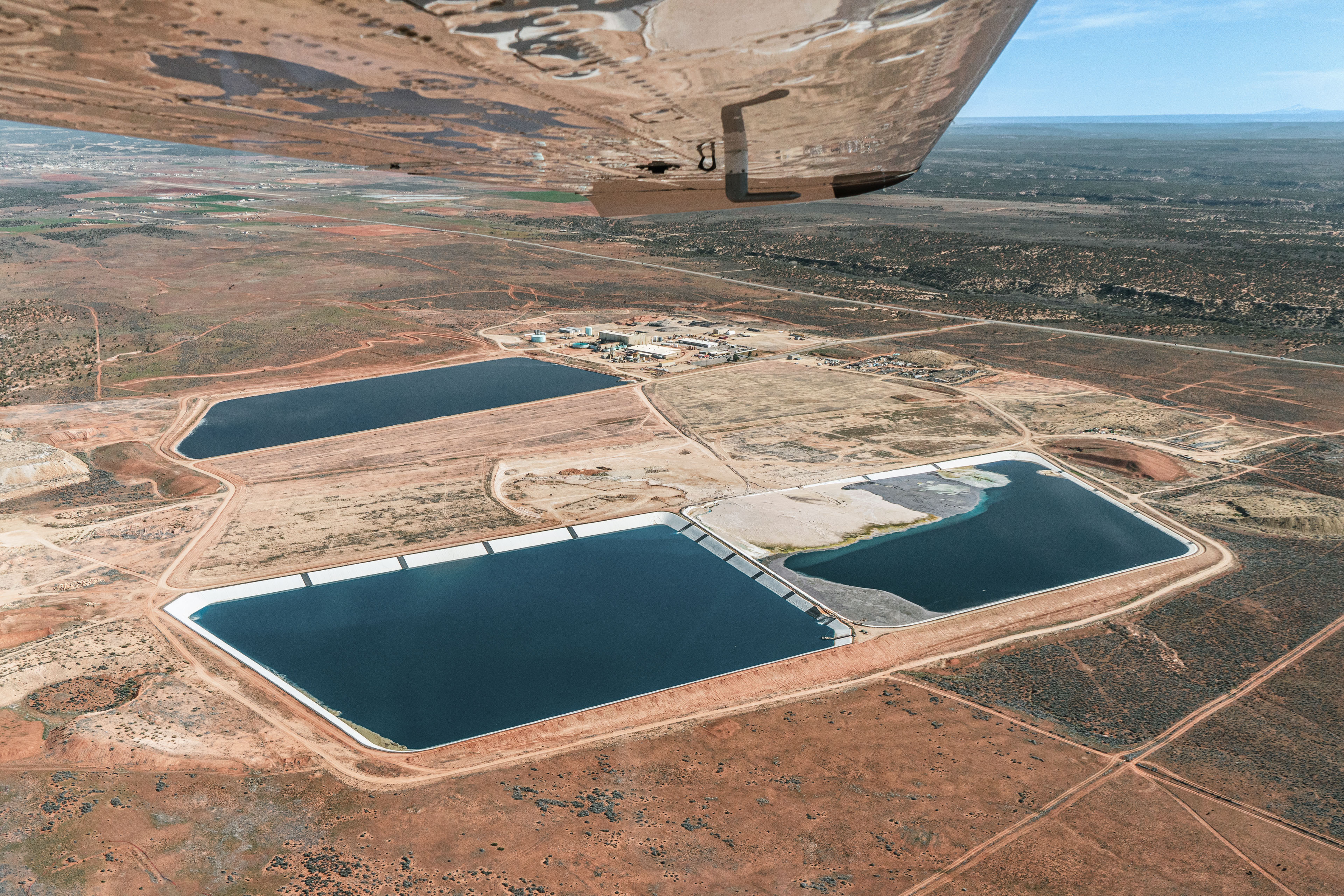 An aerial view shows tailing ponds amidst a mesa landscape. The wing of an airplane from which the photo was taken is seen in the frame.