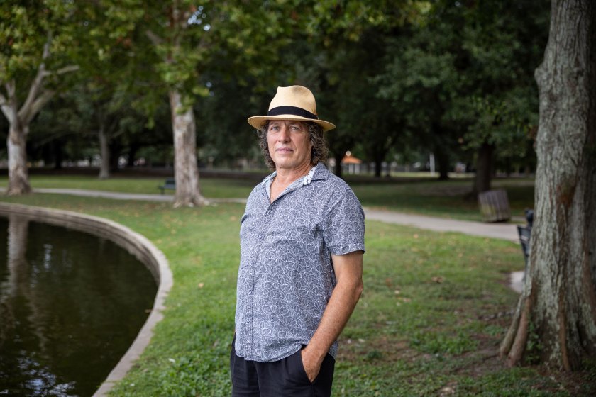 A man in a beige brimmed hat and patterned blue short-sleeve shirt stands in a park near a lake and looks at the camera.