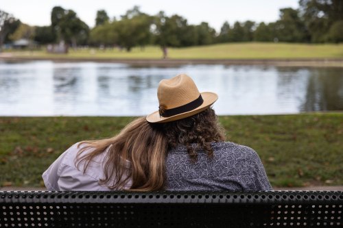 A view from behind of a man and teenage girl sitting on a park bench and looking out at a lake. The girl rests her head on the man's shoulder.