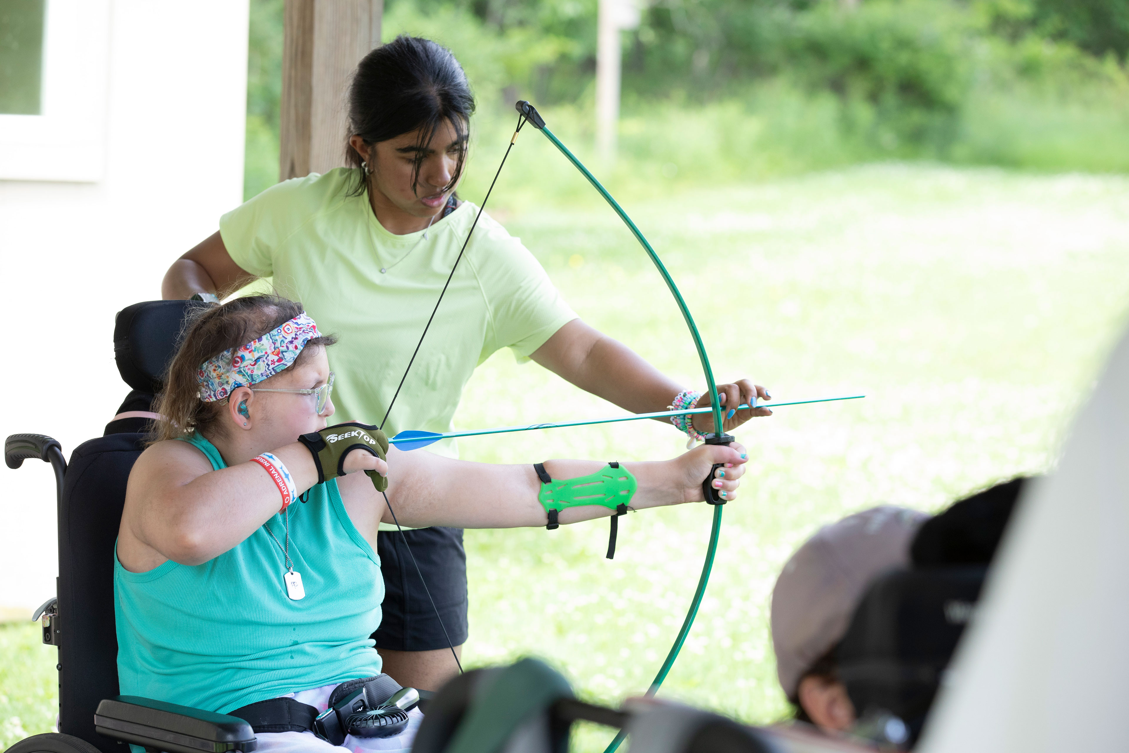A girl in a wheelchair pulls an arrow against a bow while a young woman standing behind her helps position the arrow's aim.