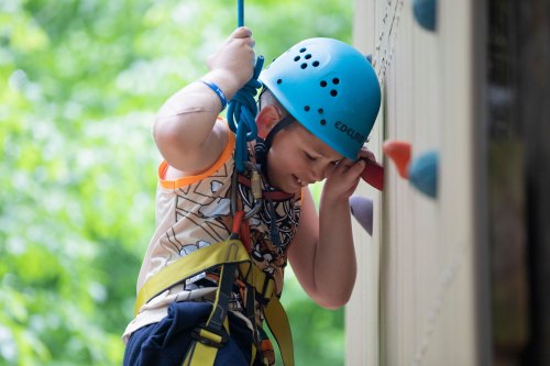 A boy wearing a blue helmet and climbing harness smiles while suspended from a rock climbing wall.