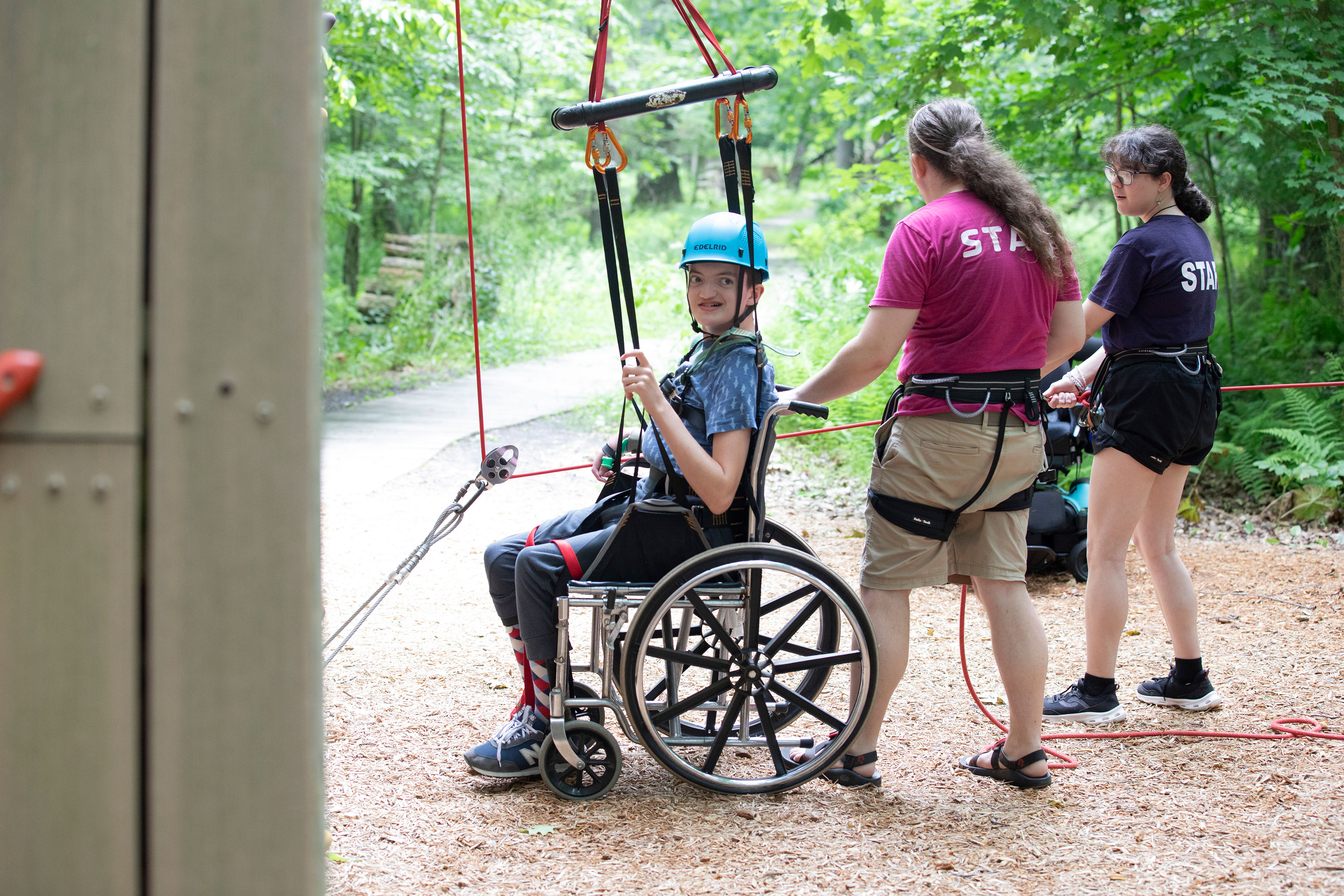 A boy in a blue helmet sits in a wheelchair in front of a rock climbing wall as two other people prepare ropes leading to the harness the boy is wearing.
