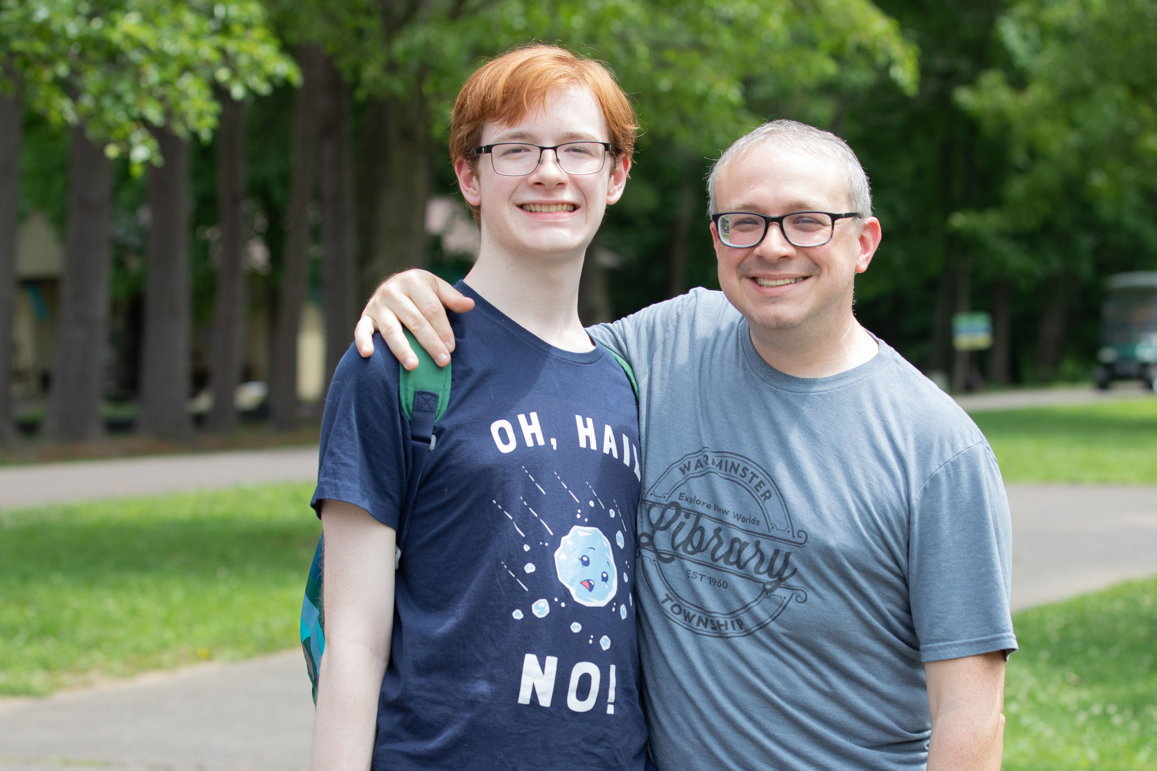 A man stands outside with his arm around the shoulders of his teenage son. Both smile at the camera.