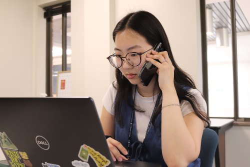 A woman holding a phone to her left ear sits in front of a laptop and looks at the screen.