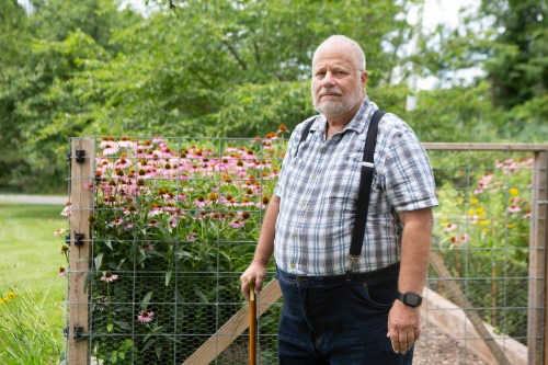 An older man, holding a cane and wearing a button down plaid shirt with suspenders, stands in front of a blooming garden.